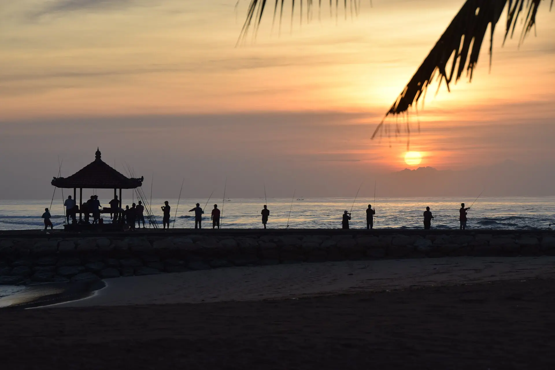 Sunset over the sea at Nusa Dua, with silhouettes of people beside a traditional Balinese pavilion